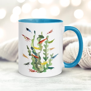 May include: A white ceramic mug with a bright blue handle and interior rim. The exterior features a watercolour illustration of green aquatic plants surrounded by various small, colourful freshwater shrimp in shades of red, blue, yellow, and black.