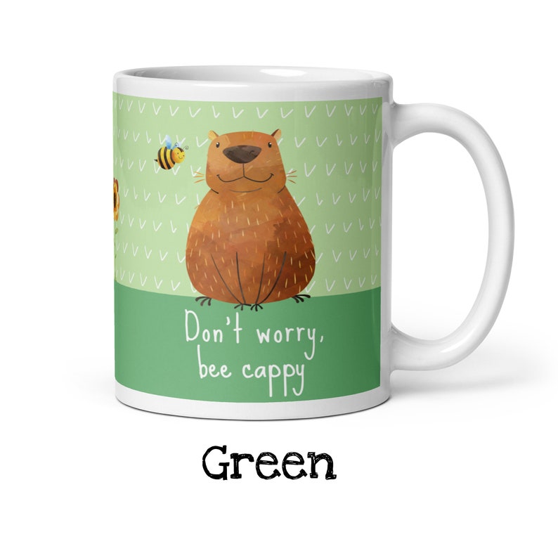 Op de afbeelding: Een witte keramische mok met een groene achtergrond en een cartoonillustratie van een bruine beer met een bij die erboven vliegt. De tekst "Don't worry, bee cappy" is in zwart op de mok gedrukt.