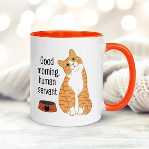 May include: White ceramic mug with an orange rim and handle. The mug features a cartoon orange tabby cat with green eyes and the text "Good morning, human servant".