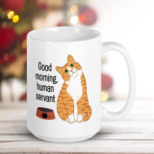 A white ceramic mug depicting an orange tabby cat waiting expectantly by its empty food bowl, with the text "Good morning, human servant" printed above the bowl.