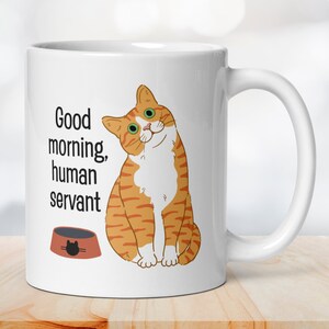 A white ceramic mug depicting an orange tabby cat waiting expectantly by its empty food bowl, with the text "Good morning, human servant" printed above the bowl.