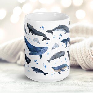May include: White ceramic mug with a watercolor pattern of various whales and dolphins in shades of blue and gray. The mug features a repeating pattern of the sea creatures against a white background.