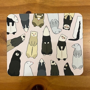 May include: A rectangular mouse mat with a repeating pattern of cartoon ferrets and skunks in various poses on a light pink background. The animals are depicted in black, white, brown, and tan colours.