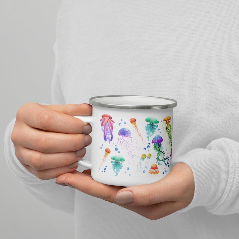 White enamel jellyfish mug cradled in hands showing the reverse side with colourful illustrated jellyfish species in green, purple, orange and red with blue bubbles — a coastal camping gift.