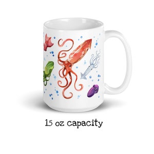 May include: White ceramic mug featuring watercolor illustrations of various sea creatures, including squid and octopus, in shades of red, green, and purple. The mug has a 15 oz capacity.