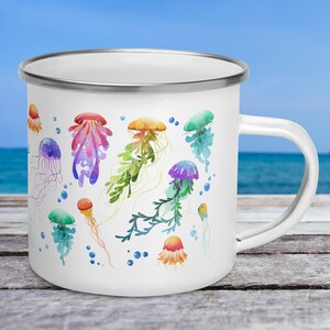 A 12-ounce enamel mug rests on an outdoor table with the ocean behind it. The mug has a silver rim and its handle is on the right. Printed on the white mug are colourful watercolour illustrations of jellyfish in pink, green, orange, purple and blue.