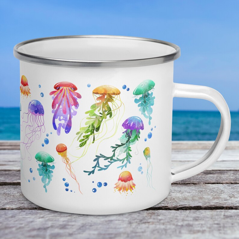 A 12-ounce enamel mug rests on an outdoor table with the ocean behind it. The mug has a silver rim and its handle is on the right. Printed on the white mug are colourful watercolour illustrations of jellyfish in pink, green, orange, purple and blue.