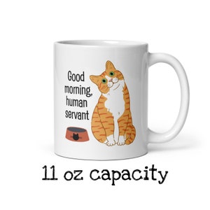 May include: White ceramic mug with a cartoon orange tabby cat with green eyes. The cat is sitting and looking to the right. The mug has the text "Good morning, human servant" and a cartoon image of a cat food bowl. The mug has a 11 oz capacity.