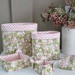 May include: A collection of fabric storage baskets and containers in different sizes. The baskets have a green floral pattern with white flowers and a pink polka dot trim. The containers are displayed on a white surface.