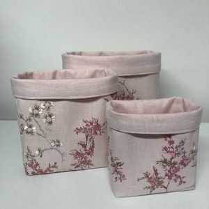May include: Three pink fabric storage baskets with floral embroidery. The baskets are different sizes and are arranged in a row from largest to smallest.