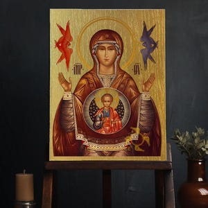 Könnte beinhalten: Eine religiöse Ikone, die die Jungfrau Maria mit dem Jesuskind zeigt. Die Ikone ist auf einem Holzpaneel mit goldenem Hintergrund gemalt und hat ein rot-blaues Farbschema. Die Ikone ist in einem goldenen Rahmen gefasst und auf einem Holzständer ausgestellt.