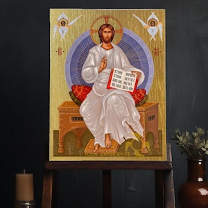 Puede incluir: Un icono religioso que representa a Jesucristo sentado en un trono, sosteniendo un libro con texto en un idioma extranjero. El icono está pintado en un estilo tradicional con acentos dorados y un fondo azul. El icono está enmarcado y se muestra en un caballete de madera.