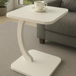 Puede incluir: Mesa auxiliar blanca en forma de C con tapa y base cuadradas. Una taza y un platillo blancos con la palabra "NEZO" están sobre la mesa. La mesa está sobre una alfombra gris junto a un sofá gris.