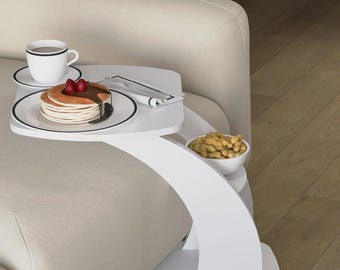 Op de afbeelding: Een witte, gebogen bijzettafel met een dienblad met pannenkoeken met siroop en aardbeien, een kopje koffie en een kleine kom pinda's. De tafel staat naast een beige bank.