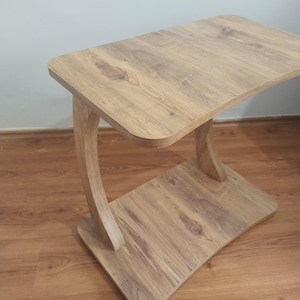 May include: A light brown wooden side table with a curved leg design. The table has a rectangular top and a lower shelf, both with a wood grain pattern. The table measures approximately 61 cm long, 30 cm wide, and 51 cm high.
