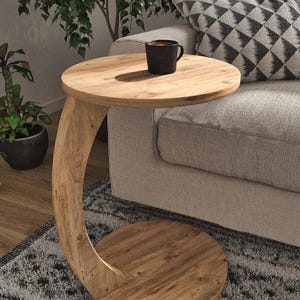 Puede incluir: Una mesa auxiliar de madera marrón claro con una parte superior y una base circulares, conectadas por un soporte curvo. Una taza negra está sobre la superficie de la mesa. La mesa está junto a un sofá gris y un cojín estampado.