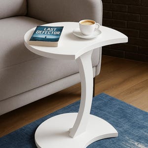 May include: White, curved side table with a book titled "LAST DEFECTOR" and a cup of coffee on top. The table has a crescent-shaped base and a curved support. It sits on a blue rug next to a gray sofa.
