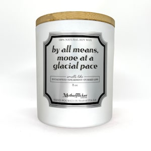 May include: A white candle with a wooden lid. The label reads "by all means, move at a glacial pace" and lists scents like eucalyptus and spearmint. The candle is 8 oz.