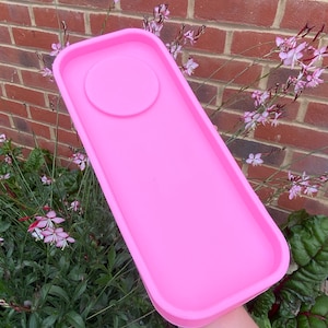 Puede incluir: Una bandeja rectangular rosa con una hendidura redonda en el centro. La bandeja está hecha de un material liso y brillante.