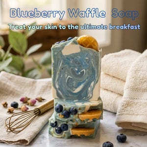 May include: A stack of three rectangular bars of soap with a blue and white marbled pattern, topped with small golden waffle-shaped pieces and blueberries. The text reads Blueberry Waffle Soap, Treat your skin to the ultimate breakfast, BloomedBubblesSoaps.Etsy.com.