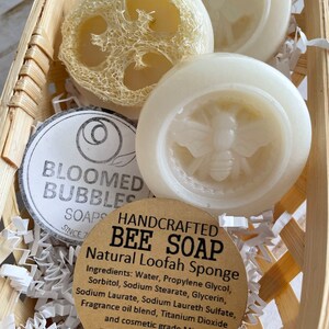May include: Three handmade bee soap bars in a wicker basket with shredded paper. The soap bars are white and feature a bee design. One soap bar is a natural loofah sponge. The label reads "HANDCRAFTED BEE SOAP Natural Loofah Sponge" and lists the ingredients. The net weight is 1.1 oz.