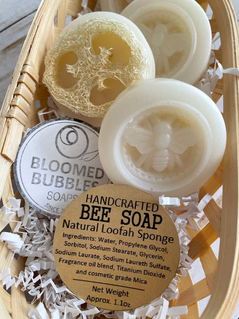 May include: Three handmade bee soap bars in a wicker basket with shredded paper. The soap bars are white and feature a bee design. One soap bar is a natural loofah sponge. The label reads "HANDCRAFTED BEE SOAP Natural Loofah Sponge" and lists the ingredients. The net weight is 1.1 oz.