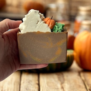 Può includere: Una saponetta marrone con una parte superiore decorativa con panna montata bianca, una piccola zucca arancione e foglie verdi. Il sapone ha vortici arancioni e gialli.