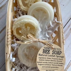 May include: Three round bars of natural loofah soap in a woven basket. The soap is white with a loofah pattern. The label reads "HAND CRAFTED BEE SOAP Natural Loofah Sponge" and lists the ingredients. The net weight is 1.1 oz.