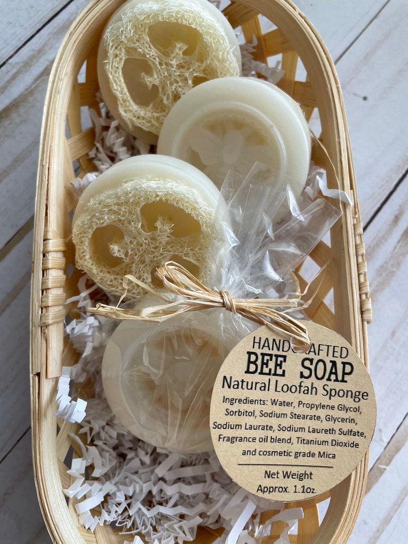 May include: Three round bars of natural loofah soap in a woven basket. The soap is white with a loofah pattern. The label reads "HAND CRAFTED BEE SOAP Natural Loofah Sponge" and lists the ingredients. The net weight is 1.1 oz.