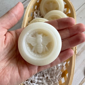 May include: A close-up of a white, round soap bar with a bee design. The soap is in a hand and is surrounded by white shredded paper in a woven basket.