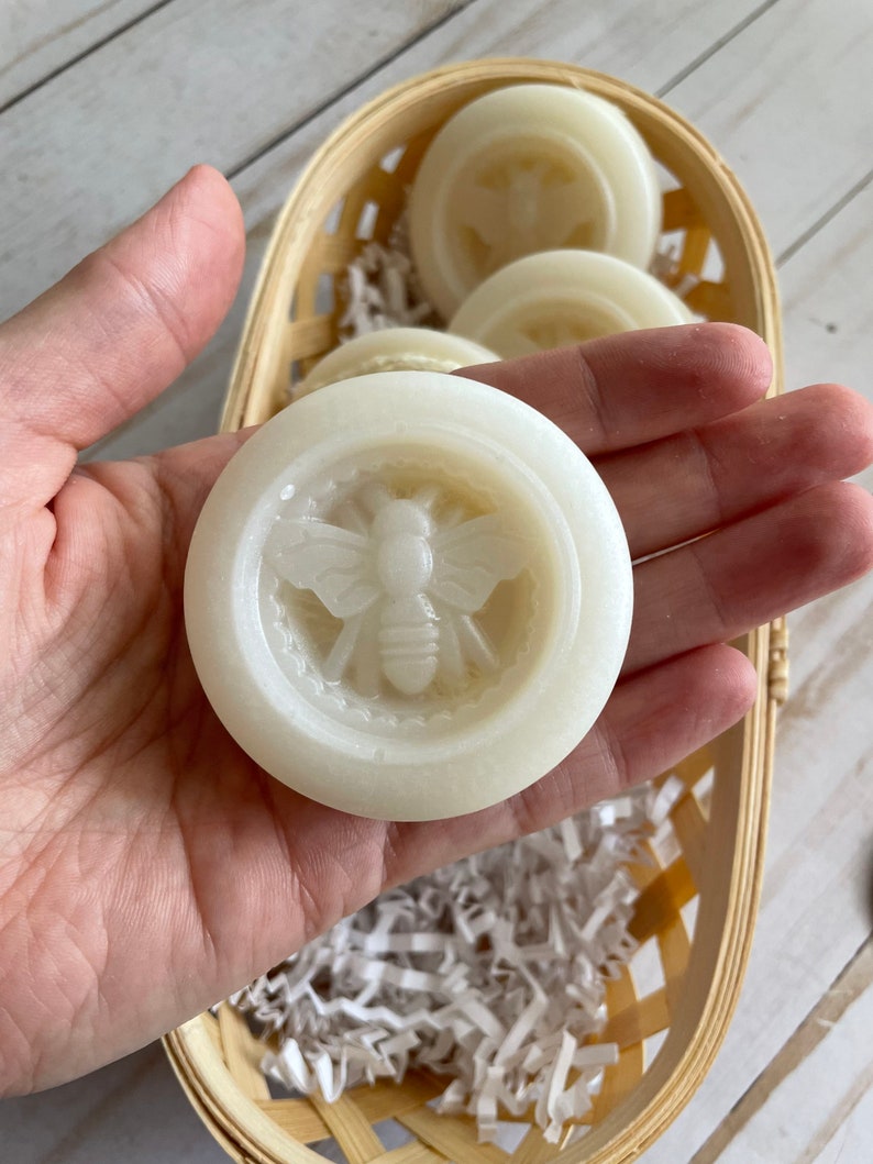 May include: A close-up of a white, round soap bar with a bee design. The soap is in a hand and is surrounded by white shredded paper in a woven basket.