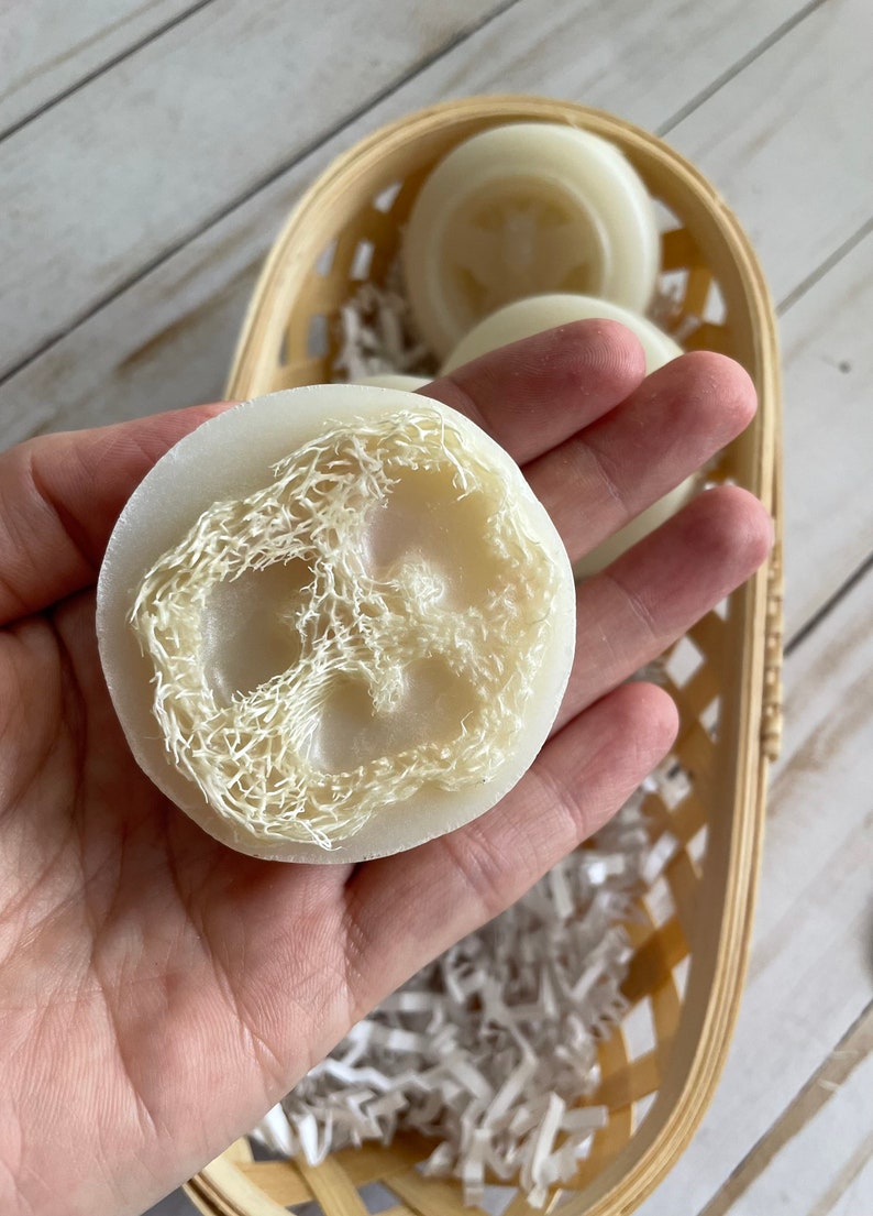 May include: A round, white soap bar with a loofah embedded in the centre. The soap is being held in a hand, and there are other soap bars in a basket in the background.