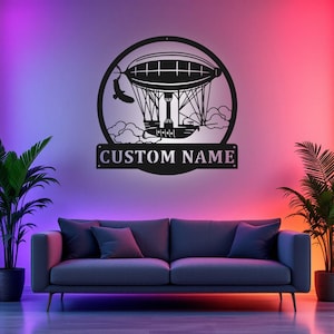 Puede incluir: Arte de pared de metal negro con un globo aerostático con un pájaro volando por encima. El arte está en un círculo con las palabras "CUSTOM NAME" debajo.