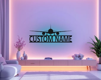 Puede incluir: Una silueta de avión de metal negro con el texto "CUSTOM NAME" debajo. El avión está centrado en una pared rosa claro con un degradado azul claro. La pared está iluminada con luces blancas cálidas.