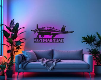 Puede incluir: Silueta negra de metal de un avión pequeño con el texto "CUSTOM NAME" debajo. El avión está montado en una pared en una habitación con un sofá y plantas.
