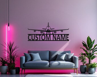 Puede incluir: Decoración de pared de metal negro en forma de avión con el texto "CUSTOM NAME" debajo. La decoración está montada en una pared rosa sobre un sofá gris.