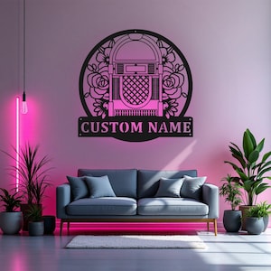 May include: Black metal jukebox wall art with a floral design and the text "CUSTOM NAME". The jukebox is set inside a circular frame with roses. The wall art is mounted on a pink wall above a gray sofa.