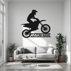 May include: Black metal silhouette of a motocross rider jumping a dirt bike. The silhouette is mounted on a white wall above a gray couch. The text "CUSTOM NAME" is below the silhouette.