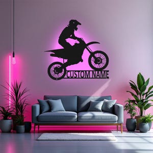 May include: Black metal silhouette of a motocross rider on a dirt bike with the text "CUSTOM NAME" below. The silhouette is mounted on a wall in a room with a couch and plants.