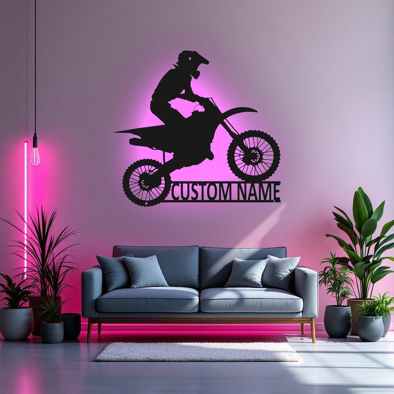 May include: Black metal silhouette of a motocross rider on a dirt bike with the text "CUSTOM NAME" below. The silhouette is mounted on a wall in a room with a couch and plants.
