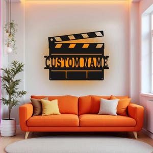 Può includere: Decorazione da parete in metallo nero a forma di ciak con il testo "CUSTOM NAME" in arancione. Il ciak è montato su una parete bianca sopra un divano arancione.