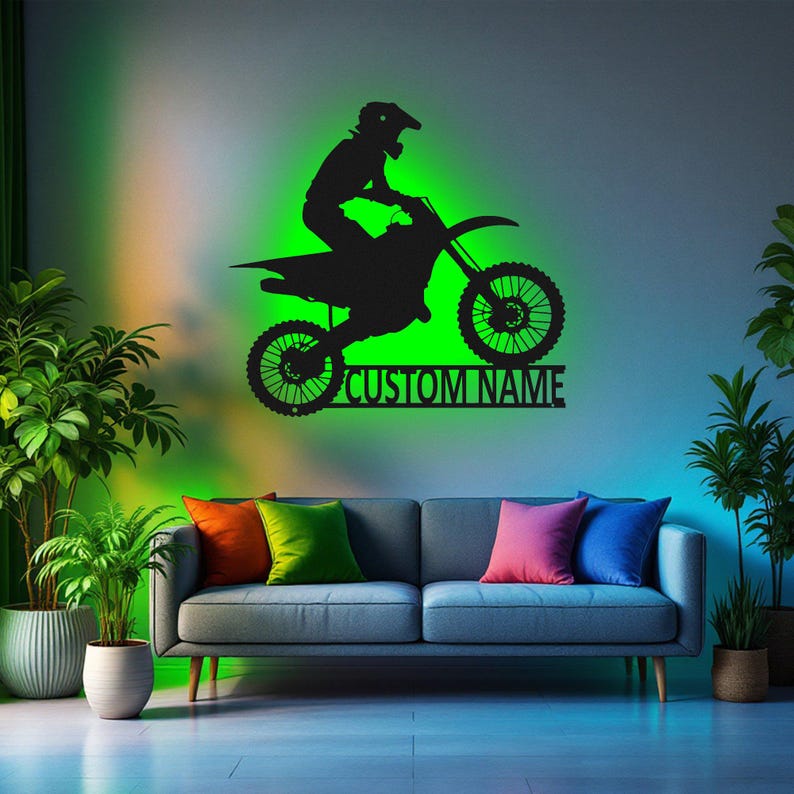 May include: Black silhouette of a motocross rider on a dirt bike with the text "CUSTOM NAME" below. The image is on a wall with a green and orange gradient behind it. A couch with pillows is in the foreground.