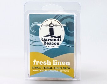 May include: A small batch soy wax candle in a clear plastic package. The candle is labeled "fresh linen" and has a light blue and yellow design with a lighthouse graphic. The label also says "lemon-floral-light musk" and "Garsnett Beacon Candle Co."