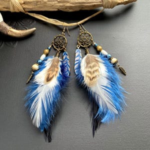 Può includere: Un paio di orecchini fatti a mano con un design acchiappasogni. Gli orecchini presentano piume blu, bianche e marroni, impreziosite da perline e piccoli ciondoli in metallo. Gli orecchini sono sospesi a ganci color oro.