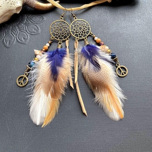 Op de afbeelding: Een paar hangende oorbellen met bronzen dromenvangerbedels, kleine veelkleurige kralen en vredestekenhangers. Elke oorbel is geaccentueerd met lange, zachte veren in paarse, bruine en witte tinten, hangend aan dunne beige suède koordjes.