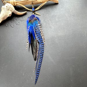 Könnte beinhalten: Ein langer Ohrring mit einem Bündel aus leuchtend blauen und schwarzen Federn mit Streifenmuster. Der Ohrring ist mit einer kleinen blauen Perle, einem metallischen Federanhänger und einem Hakenverschluss versehen, was für eine geschichtete Optik sorgt.