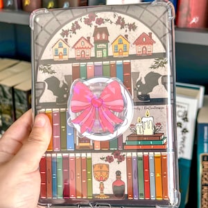 Peut inclure: Coque de tablette transparente avec un motif de bibliothèque. La coque présente un support de téléphone en forme de nœud rose, des maisons miniatures et des illustrations de livres, de bougies et d'un calice. La coque protège la tablette.