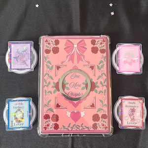 Puede incluir: Una funda de teléfono rosa con un lazo y las palabras "One More Chapter" está rodeada de cuatro agarres de teléfono a juego. Cada agarre presenta un diseño diferente y la palabra "Lover" con las frases "Fanma", "Gothie Fantasy", "Dark Romance" y un diseño de fresa.