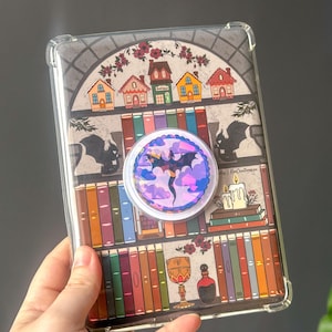Könnte beinhalten: Transparente Handyhülle mit einer farbenfrohen Illustration eines Bücherregals, Häusern und einem Drachen. Ein Pop-Socket mit Drachendesign ist angebracht. Die Hülle hat einen transparenten Rand.