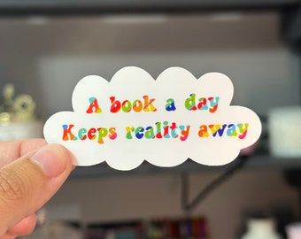Puede incluir: Una pegatina blanca en forma de nube con el texto "A book a day keeps reality away" en colores arcoíris.